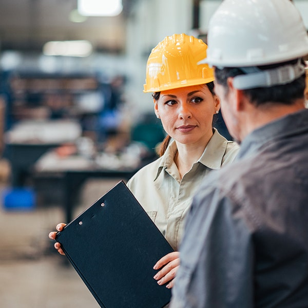 Woman with a clipboard and yellow hard hat listening intently to her male coworker