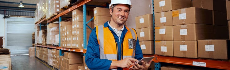 Male associate smiling in a warehouse in his uniform of a hardhat and a bright orange vest