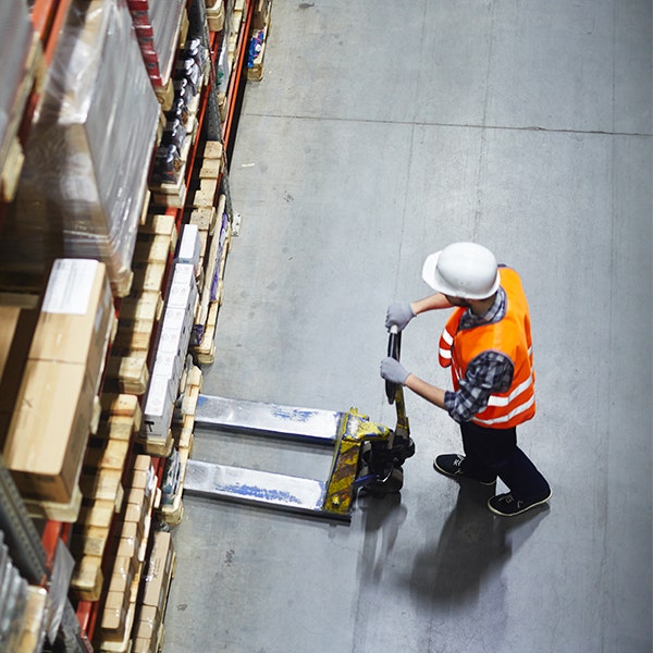 A man wearing a hardhat with an orange vest while using a pallet jack to lift up pallets in a warehouse